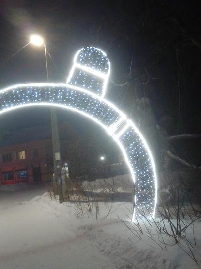 Фото арки с гирляндой, которая светится белым светом. Из верхней части арки выпирает небольшая, но явно фаллическая фигура.

На улице вечер, темно. Вокруг снег. Светит фонарь.